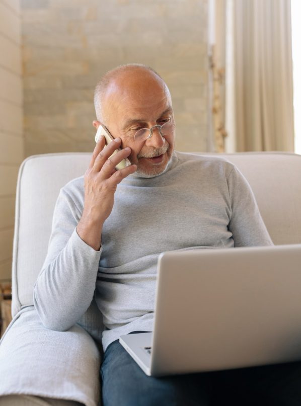 An older man talks on his cell phone while looking at his laptop